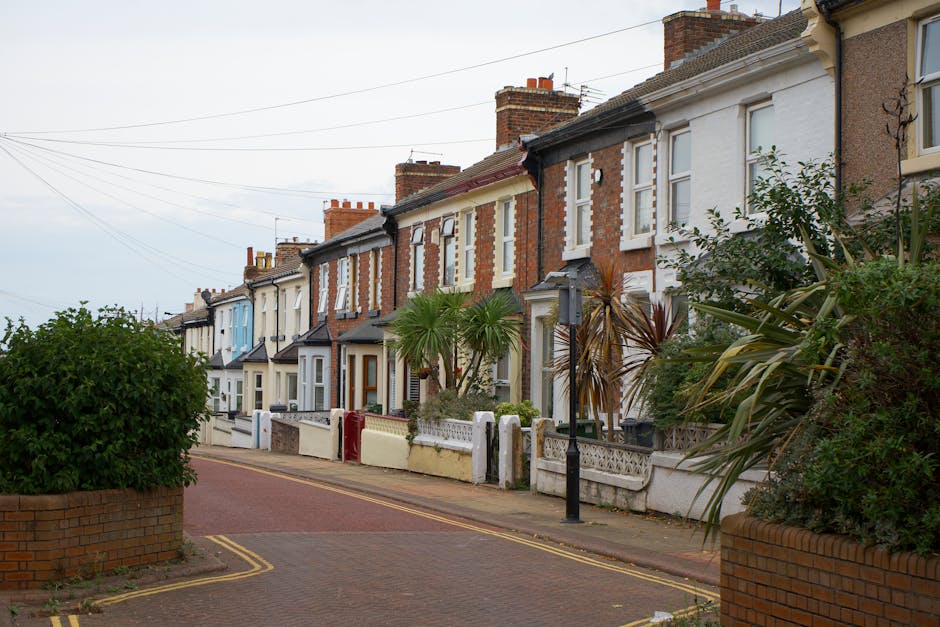 A quiet residential street featuring a row of traditional terraced houses with varying brick facades, some painted in pastel shades, and white window frames. The front gardens are neatly maintained, w